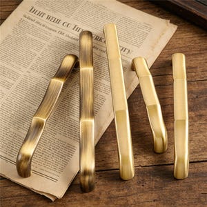 Puede incluir: Un conjunto de tiradores de armario en tono latón de varias formas. Algunos son curvos, otros rectos. El herraje se dispone sobre un periódico antiguo y una superficie de madera, mostrando su diseño y acabado.