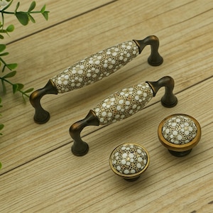 Puede incluir: Juego de herrajes decorativos para armarios con dos tiradores largos y dos pomos. Los tiradores y pomos tienen un estampado floral crema sobre fondo marrón, con detalles metálicos color bronce. Para armarios y cajones.