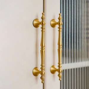 May include: Two ornate, gold-colored door handles. Each handle has a long, slender body with decorative detailing and a rounded base. The handles are mounted on a white surface, suggesting a cabinet or door.