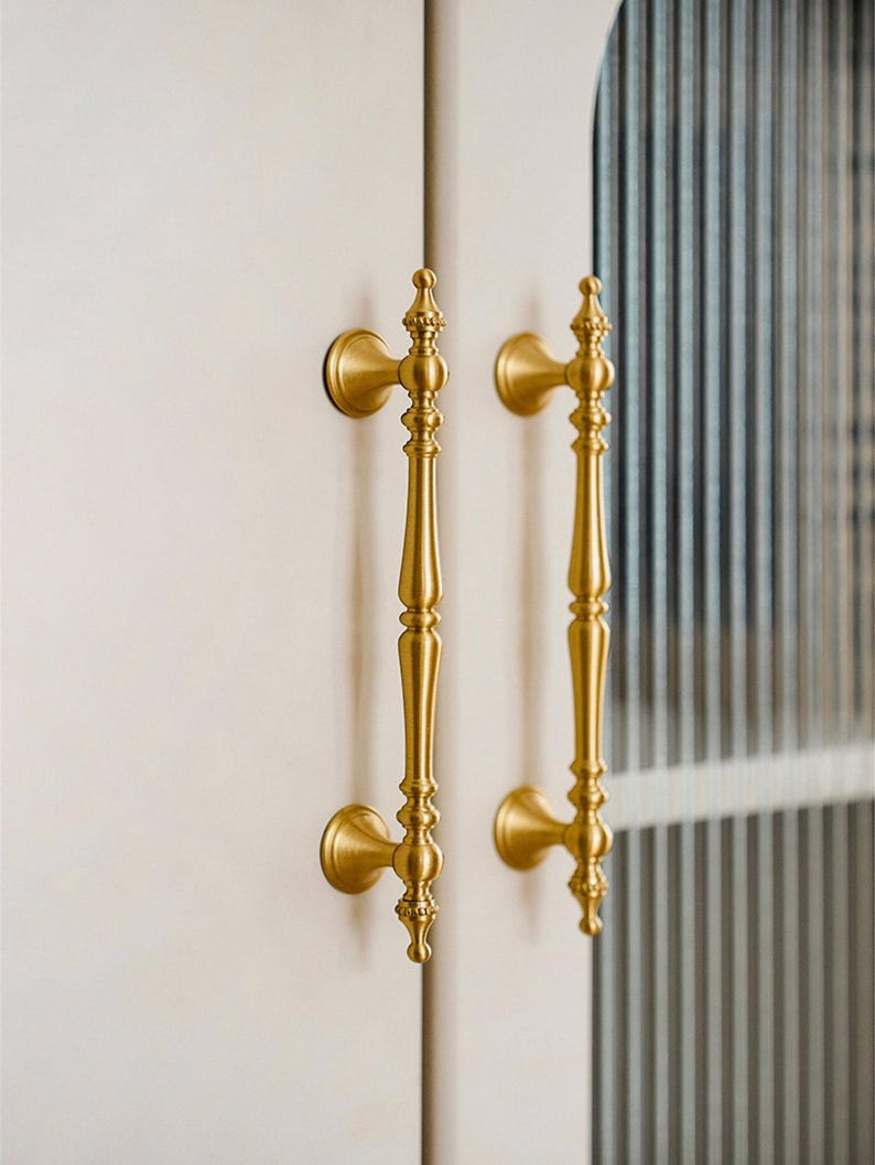 May include: Two ornate, gold-colored door handles. Each handle has a long, slender body with decorative detailing and a rounded base. The handles are mounted on a white surface, suggesting a cabinet or door.