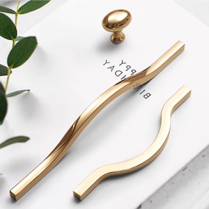 Pode incluir: Um conjunto de ferragens douradas para armários, incluindo duas puxadores curvos e um puxador. Os puxadores têm um design elegante e moderno, e o puxador é oval. O hardware é exibido em uma superfície branca com o texto "HAPPY BIRTHDAY" visível.