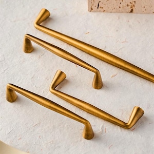 May include: Four antique brass cabinet pulls in varying sizes are arranged on a textured, off-white surface. The pulls have a curved design with a cylindrical base and a straight handle. The warm, golden colour of the hardware adds a touch of elegance.