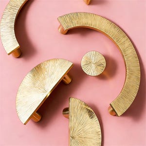 Könnte beinhalten: Goldfarbene Schrankbeschläge auf rosa Hintergrund. Das Set umfasst halbkreisförmige und runde Knöpfe mit einer strukturierten, sonnenstrahlenartigen Oberfläche. Geeignet für Möbel oder Schränke.