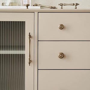 以下が含まれることがあります： アンティーク真鍮金具のキャビネットのクローズアップ。キャビネットはガラスパネルのドアと丸いノブ付きの引き出しを備えています。いくつかの真鍮製のハンドルとノブが見え、ヴィンテージ感を演出しています。