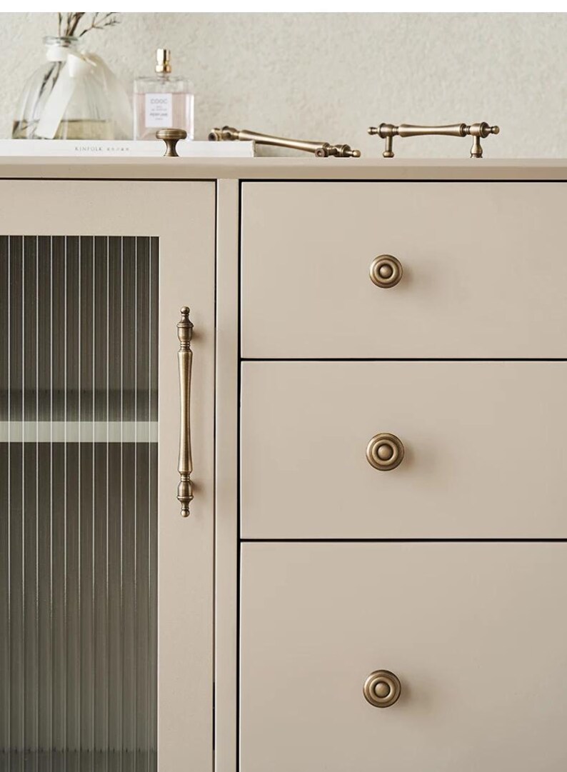 以下が含まれることがあります： アンティーク真鍮金具のキャビネットのクローズアップ。キャビネットはガラスパネルのドアと丸いノブ付きの引き出しを備えています。いくつかの真鍮製のハンドルとノブが見え、ヴィンテージ感を演出しています。