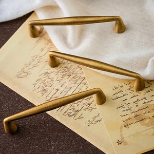 Puede incluir: Tres tiradores de armario de latón antiguo con un diseño curvo. Los tiradores están dispuestos sobre un fondo de papel vintage con escritura manuscrita y un paño blanco. La ferretería tiene un tono dorado cálido.