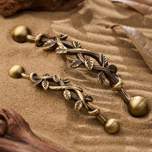 Peut inclure: Deux poignées de meuble en bronze antique avec un motif de feuilles et de vigne. Chaque poignée comporte un bouton rond à chaque extrémité et un centre décoratif avec des feuilles et des vignes détaillées. Les poignées sont posées sur une surface sablonneuse.