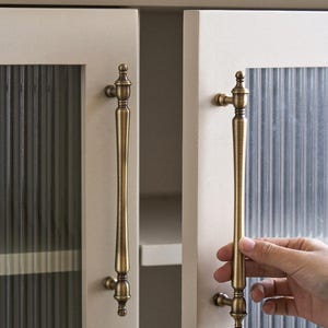 以下が含まれることがあります： クリーム色のキャビネットドア、リブ付きガラスパネルとアンティーク真鍮のハンドル。ハンドルは長く装飾的で、上部と下部に装飾的なディテールがあります。キャビネットドアは明るいクリーム色です。