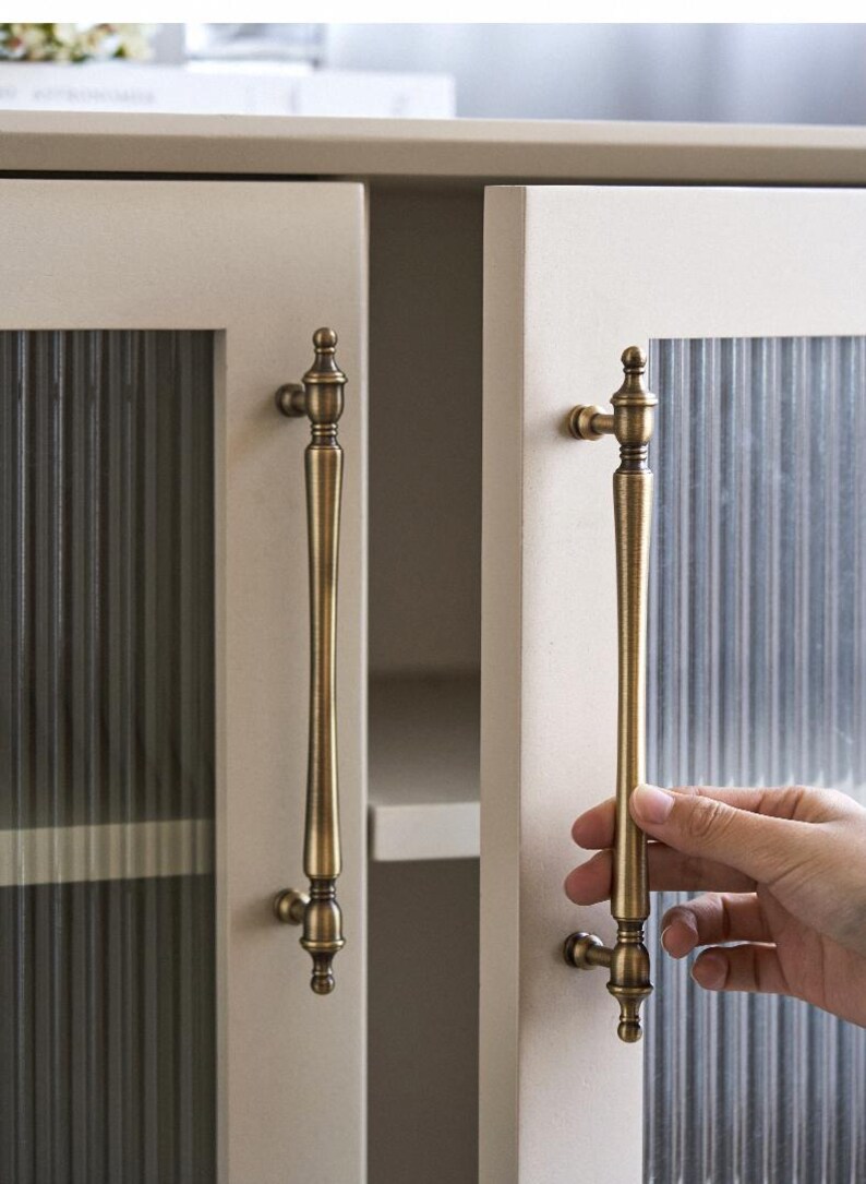 以下が含まれることがあります： クリーム色のキャビネットドア、リブ付きガラスパネルとアンティーク真鍮のハンドル。ハンドルは長く装飾的で、上部と下部に装飾的なディテールがあります。キャビネットドアは明るいクリーム色です。