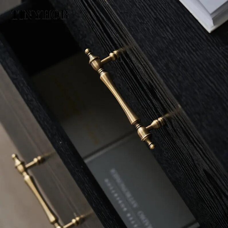 以下が含まれることがあります： 黒い木製キャビネットのクローズアップ。真鍮色の取っ手が2つ付いています。取っ手は装飾的で、曲線的なデザインです。キャビネットのドアは開いており、暗い内部と本の様子が見えます。