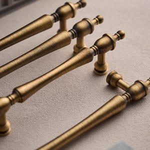 以下が含まれることがあります： アンティーク真鍮のキャビネットプル4つのクローズアップ。各プルは、端とベースに装飾的なディテールを備えた、長くテクスチャ加工されたハンドルを備えています。ハードウェアは暖かく、古くなった仕上げで、ヴィンテージまたはクラシックなスタイルを示唆しています。