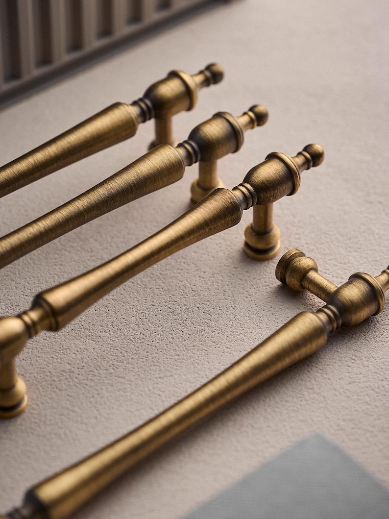 以下が含まれることがあります： アンティーク真鍮のキャビネットプル4つのクローズアップ。各プルは、端とベースに装飾的なディテールを備えた、長くテクスチャ加工されたハンドルを備えています。ハードウェアは暖かく、古くなった仕上げで、ヴィンテージまたはクラシックなスタイルを示唆しています。