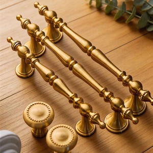 Op de afbeelding: Een verzameling goudkleurige kastbeslagen, waaronder drie lange handgrepen en twee ronde knoppen. De handgrepen zijn ongeveer 20 cm lang en de knoppen hebben een decoratieve, parelrand. De beslagen liggen op een houten oppervlak.