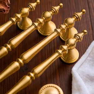 May include: Gold-colored cabinet hardware set, including several pull handles and round knobs. The handles have a detailed, ornate design with a central bar and decorative ends. The knobs feature a circular, beaded pattern. The hardware is displayed on a dark wood surface.