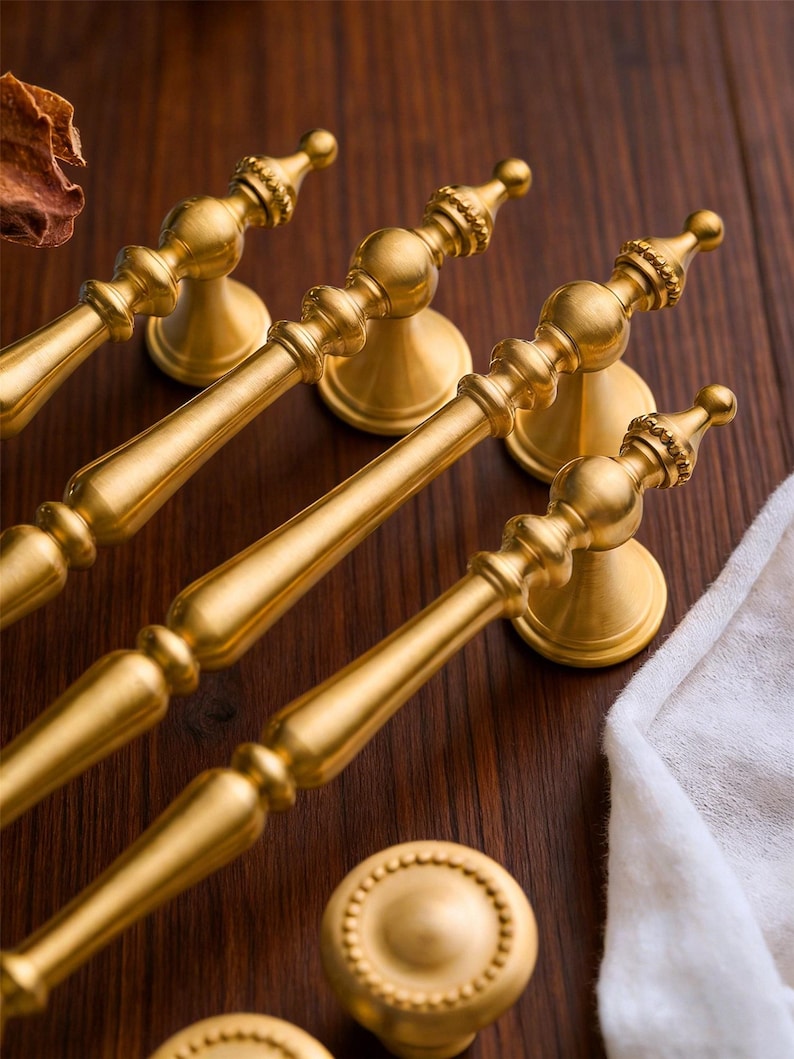 May include: Gold-colored cabinet hardware set, including several pull handles and round knobs. The handles have a detailed, ornate design with a central bar and decorative ends. The knobs feature a circular, beaded pattern. The hardware is displayed on a dark wood surface.