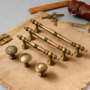 May include: An assortment of antique brass hardware, including pull handles and knobs, displayed on a burlap surface. The handles have a detailed, ornate design. The image also includes keys, a ruler, and a leather case.