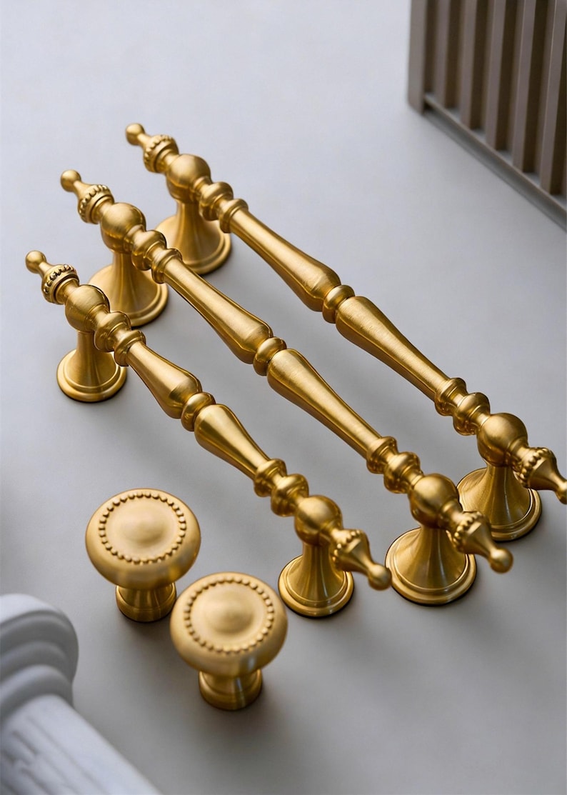 May include: A collection of gold-colored cabinet hardware, including three long pull handles and two round knobs. The handles have a detailed, ornate design, and the knobs feature a beaded edge. The hardware is arranged on a light gray surface.