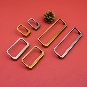 Peut inclure: Diverses poignées de meubles rectangulaires et carrées, de tons dorés et argentés. La quincaillerie est présentée sur un fond rouge, avec une pomme de pin en haut à droite. Les poignées varient en taille et en forme.