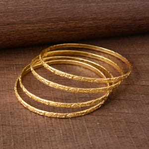 Könnte beinhalten: Ein Set aus fünf goldfarbenen Armreifen mit aufwendigen geprägten Mustern. Die Armreifen sind gestapelt, wodurch ihr detailliertes Design zur Geltung kommt. Der Schmuck wird auf einer Holzoberfläche präsentiert, die die warmen Goldtöne hervorhebt. Jeder Armreif ist etwa 0,3 cm breit.