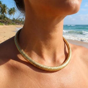 Puede incluir: Un collar dorado con un patrón texturizado en forma de escamas. El collar es circular y se coloca alrededor del cuello. El fondo muestra una playa con palmeras y olas del océano.