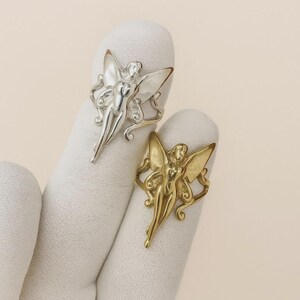 Könnte beinhalten: Zwei Ringe, einer silberfarben und einer goldfarben, jeweils mit einem Feen-Design. Die Feen haben ausgebreitete Flügel und sind in einer anmutigen Pose. Die Ringe werden auf einer weißen Oberfläche präsentiert.