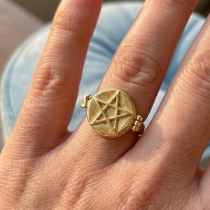 Peut inclure: Bague dorée avec un pendentif circulaire orné d'un pentagramme en relief. La bague a un design articulé et est portée au doigt. La bague est probablement en métal.