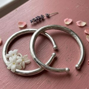 Könnte beinhalten: Zwei silberfarbene, offene Armreifen mit abgerundeten Enden sind ineinander verschlungen. Weiße Blumen und Lavendelzweige sind in der Nähe der Armreifen platziert, mit rosa Blütenblättern verstreut. Die Armreifen werden auf einer rosa Oberfläche präsentiert.