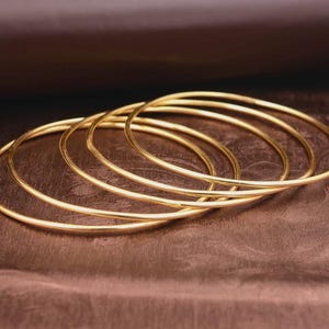 May include: Five gold-coloured bangles are displayed on a brown surface. The bangles are thin and circular, with a smooth, reflective surface. The background is a darker shade of brown, creating contrast with the bangles.