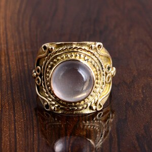 Könnte beinhalten: Ein goldfarbener Ring mit einem großen, runden, hellrosa Edelstein in der Mitte. Der Ring hat ein verziertes, strukturiertes Design um den Edelstein und am Band. Der Ring wird auf einer dunkelbraunen Oberfläche präsentiert.