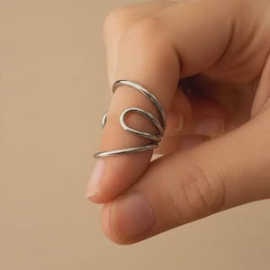 May include: A silver-colored ring with a unique, abstract design. The ring wraps around the finger with multiple curved bands, creating an open, flowing shape. The ring is made of a shiny, metallic material.