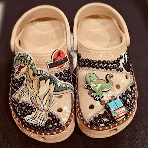 May include: A pair of beige Crocs decorated with dinosaur-themed charms and embellishments. One shoe features a Tyrannosaurus Rex patch and a Jurassic Park logo. The other shoe has a green dinosaur and a park vehicle charm, with black bead accents.