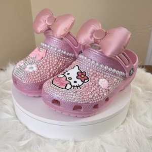 Peut inclure: Sabots roses ornés de perles et de grands nœuds en satin. Les sabots présentent un motif Hello Kitty et sont recouverts de petits ornements scintillants. Un article de chaussure décoratif et élégant.