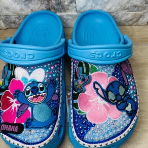 May include: A pair of blue Crocs sandals customized with Stitch from Lilo & Stitch. The sandals feature Stitch appliqués, pink and white flower designs, and are embellished with iridescent beads and rhinestones. The word "OHANA" is visible on one sandal.