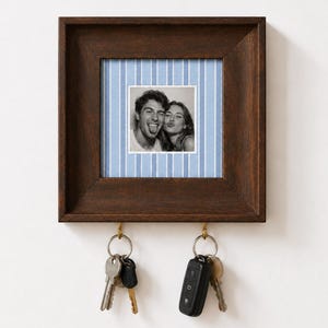 May include: A square dark brown wooden wall-mounted key holder featuring a central light blue and white pinstriped background with a black and white photograph of two people. Two brass-coloured metal hooks hang from the bottom, holding keys.