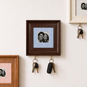 May include: A square dark brown wooden wall-mounted key holder featuring a central black and white photograph of two people. The frame surrounds a light blue and white pinstriped fabric background. Two gold metal hooks hold sets of keys.