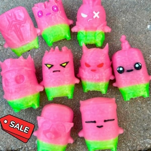 May include: A collection of nine small, pink and green plastic toy figures with various facial expressions, including some with X-shaped eyes, pointed ears, or simple dots for eyes, arranged together with a red tag labeled SALE.