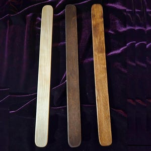 May include: Three wooden craft sticks in varying shades of brown and tan are arranged vertically against a dark purple velvet background. The sticks are rectangular with rounded ends, showcasing the natural wood grain.