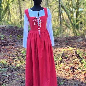 May include: A floor-length red dress with a square neckline and white lace-up front closure, worn over a white long-sleeved shirt. The dress features a fitted bodice and a full, gathered skirt, showcasing a solid red fabric with white lacing.