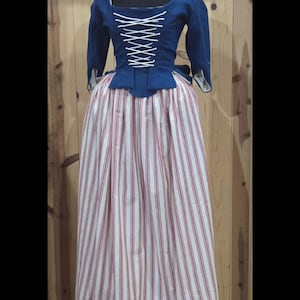 Könnte beinhalten: Ein historisches Kostüm mit einem dunkelblauen Oberteil mit weißer Schnürung und langen Ärmeln. Der Rock ist bodenlang mit roten und weißen vertikalen Streifen. Das Outfit wird auf einer schwarzen Schaufensterpuppe vor einem Holzgrund gezeigt.