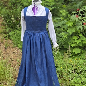 May include: A floor-length blue pinafore dress worn over a white long-sleeved shirt. The white shirt features a high collar with a ruffled edge and a front tie closure. The blue dress has a square neckline and a gathered, full-length skirt.