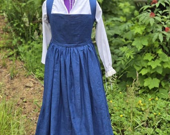 May include: A floor-length blue pinafore dress worn over a white long-sleeved shirt. The white shirt features a high collar with a ruffled edge and a front tie closure. The blue dress has a square neckline and a gathered, full-length skirt.