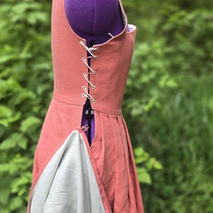 May include: A coral-colored dress form with a corset-style back, laced with white cord. The dress has a pleated skirt and a contrasting light gray lining visible at the hem. The form is set against a blurred green background.