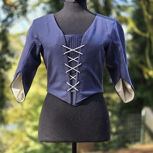 May include: A navy blue cropped jacket with a V-neck and three-quarter sleeves. The front features a white lace-up closure. The sleeves have a split design, revealing a light-colored lining. The jacket is displayed on a black mannequin.