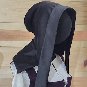 Peut inclure: Un capuchon médiéval noir avec une longue section drapée. Le capuchon est fait d'un tissu sombre et doux et fait partie d'un costume. Le capuchon est présenté sur une tête de mannequin, avec une chemise blanche et un gilet violet visible.