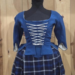 May include: A blue bodice with three-quarter length sleeves and a white lace-up front closure, paired with a dark blue and black plaid skirt. The bodice features a square neckline and a structured fit, worn over a light-colored lining visible at the cuffs.