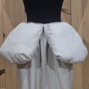 May include: A striped skirt with a unique design, featuring puffed-out sections on the hips. The fabric has a vertical stripe pattern in shades of white and gray. The skirt is displayed on a black mannequin.