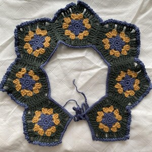 May include: A handmade crochet collar featuring a floral design. The collar is made of dark green, yellow, and blue yarn. The collar is composed of multiple hexagonal motifs joined together, with a scalloped edge and a tie closure.