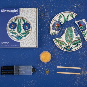 Puede incluir: Un kit Kintsugi con un plato decorativo roto con diseños florales azules. El kit incluye una caja, pegamento, polvo de oro, un pequeño gancho y palitos para remover. La caja dice "Kintsugi Kit" y "sade". El fondo es azul liso.