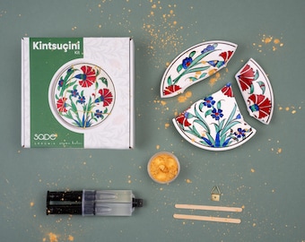 Könnte beinhalten: Ein Kintsugi-Kit mit einer Box mit der Aufschrift "Kintsugi Kit" und "Sade". Das Kit enthält einen zerbrochenen Teller mit roten und blauen Blumenmustern, Kleber, Goldpulver und Werkzeuge. Der Teller ist in drei Teile zerbrochen.