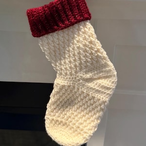 May include: A handmade Christmas stocking with a cream-colored body and a deep red cuff. The stocking is crocheted with a textured stitch, creating a cozy and festive appearance. The red cuff adds a pop of color to the design.